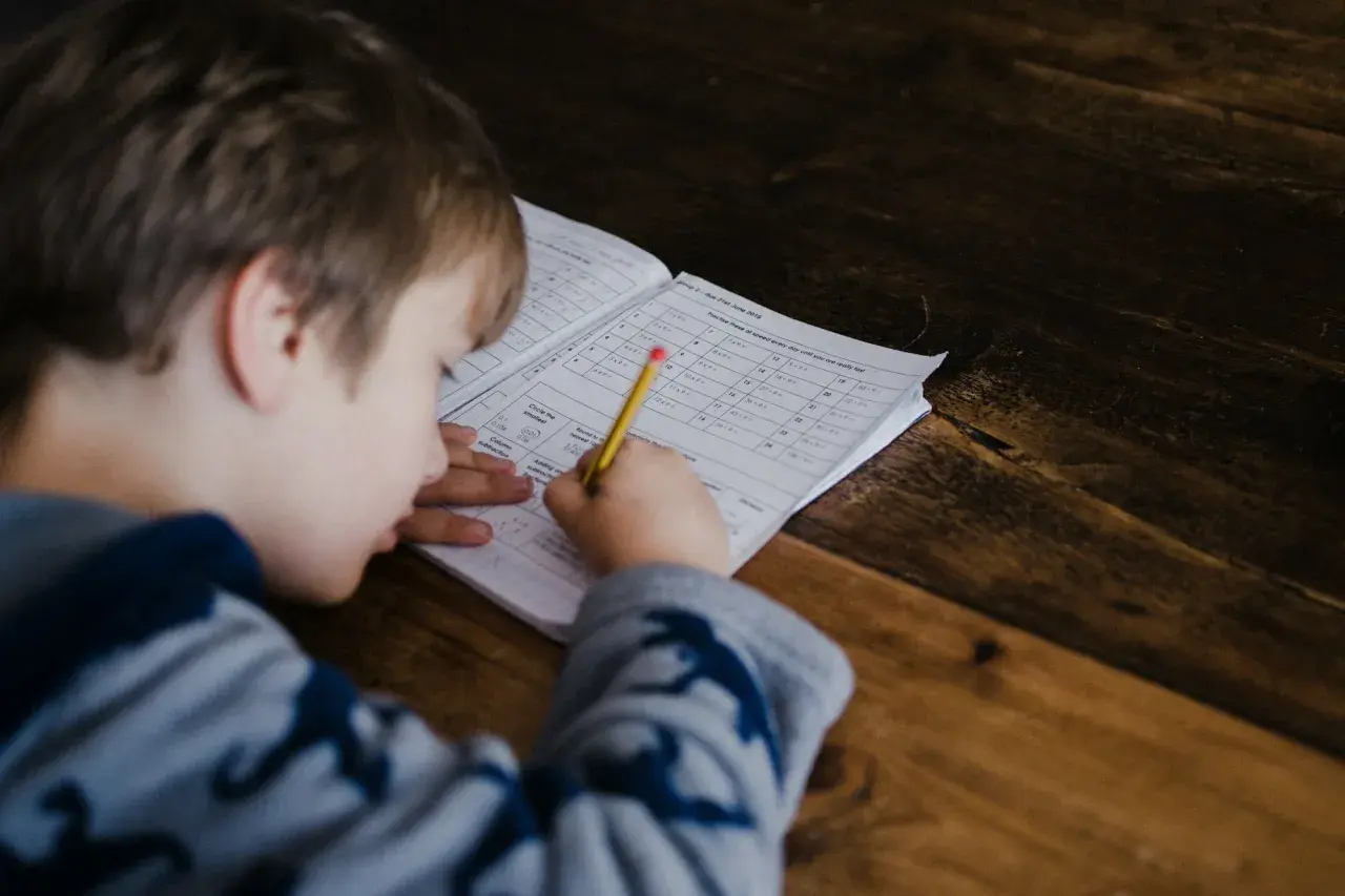 Ahşap masada matematik ödevini kurşun kalemle yapan, başını kâğıda eğmiş küçük bir erkek çocuk